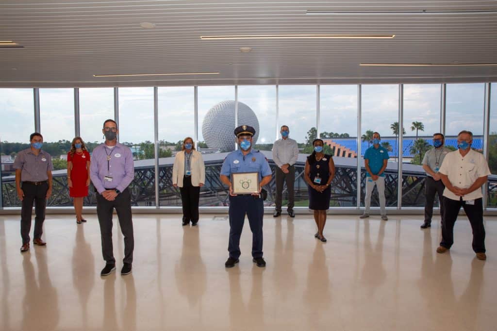 Disney cast members, all masked and socially distanced, gather indoors as one proudly displays a Disney Heroes Award certificate in the center—honoring their heroic act of saving a child.