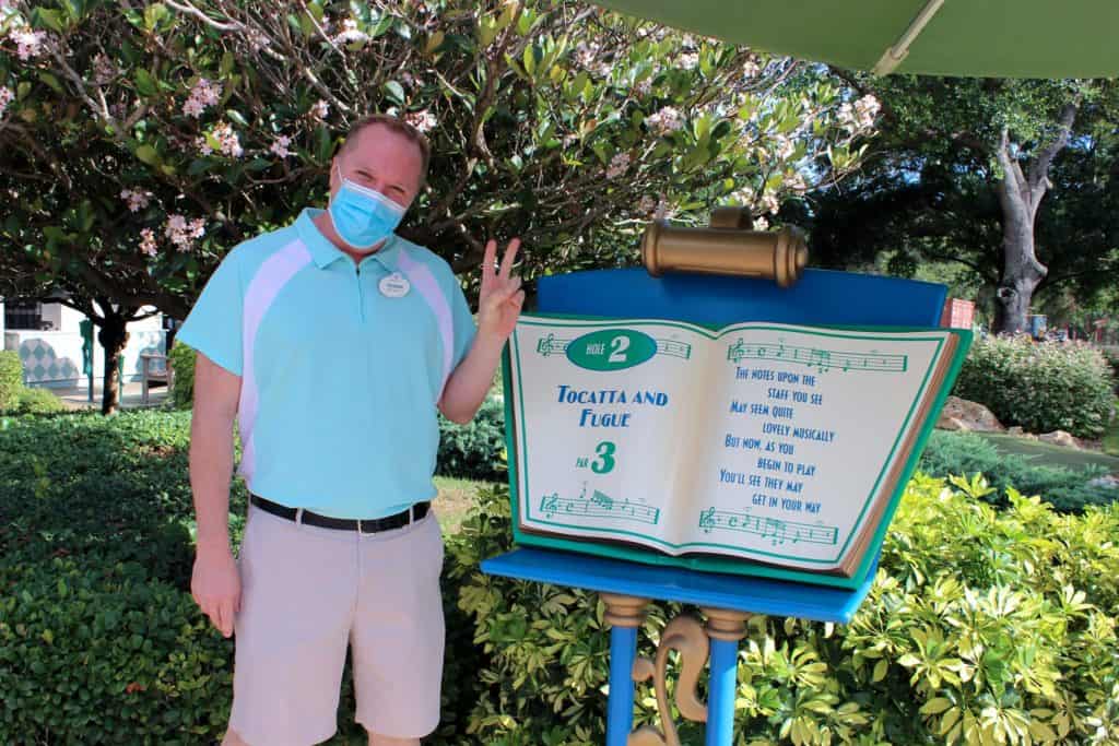 A masked guest celebrates Fantasia Gardens Miniature Golf’s 25th anniversary, flashing a peace sign beside the iconic open book sign and beaming at the camera.