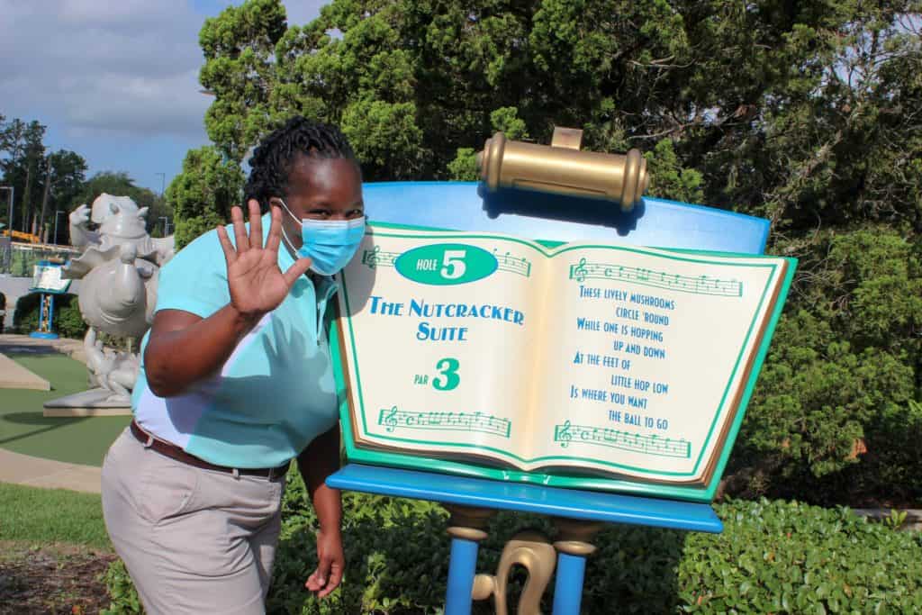 A guest sporting a blue mask greets visitors next to the Nutcracker Suite-themed mini-golf sign at Fantasia Gardens, as the beloved Walt Disney World course marks its 25th anniversary under clear, sunny skies.