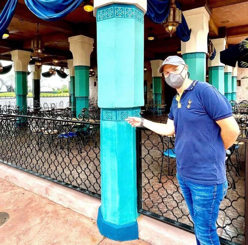 A masked guest points excitedly at a vibrant blue pillar in the open-air dining area of EPCOT’s Morocco Pavilion, surrounded by empty tables and chairs and capturing the lively atmosphere unique to this World Showcase destination.