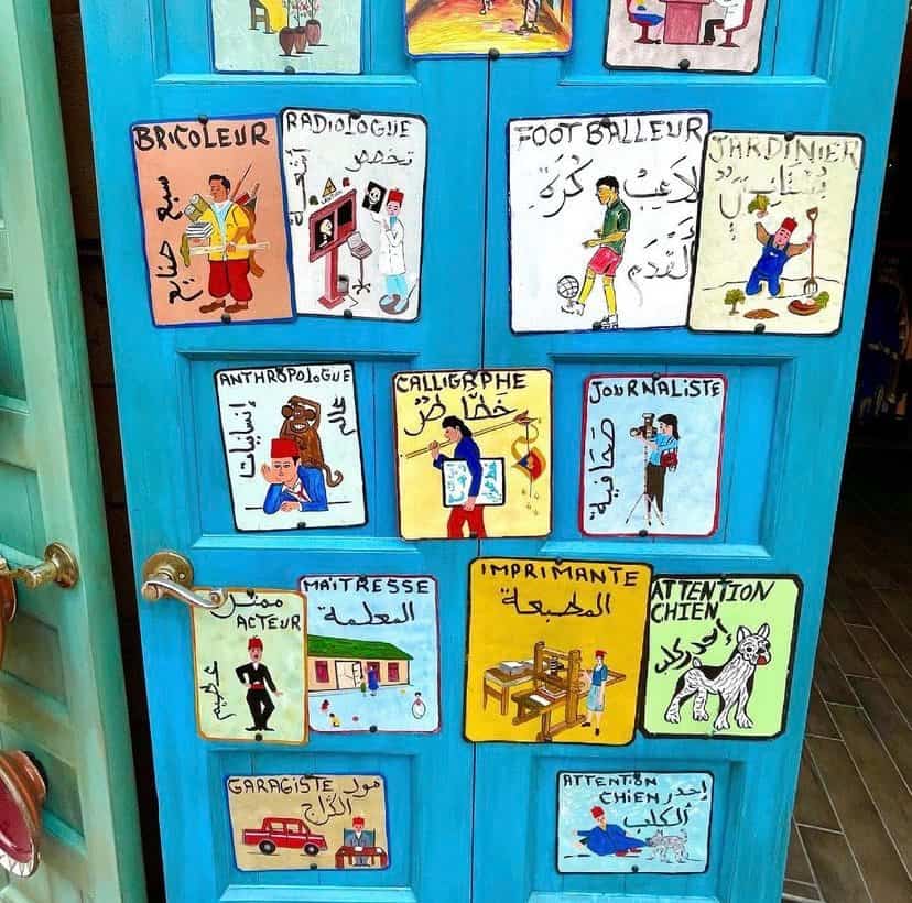 A vibrant blue door featuring lively, hand-painted job illustrations and multilingual job titles in French and Arabic on each panel, echoing the stunning artistry recently showcased at EPCOT’s Morocco Pavilion.