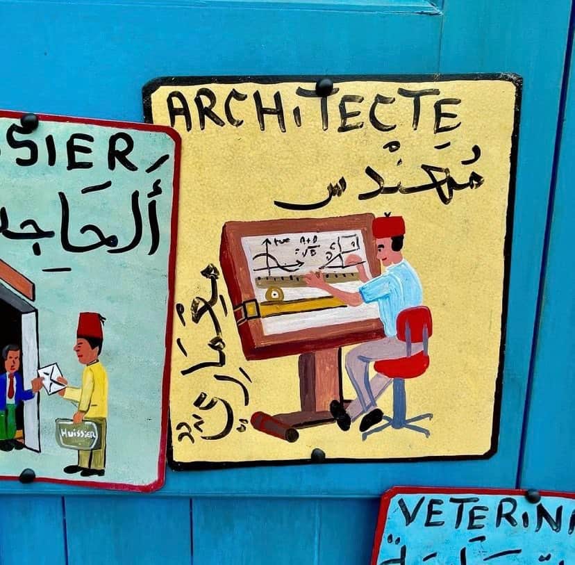 Vibrant sign featuring an architect sketching at a drafting table, topped with French and Arabic script, channeling Disney Imagineer flair and echoing the creative ambiance of EPCOT’s Morocco Pavilion.