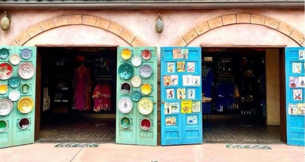 Vibrant storefronts at the EPCOT Morocco Pavilion, adorned with colorful plates and traditional art, showcase an inviting array of dresses and unique Disney souvenirs inside.