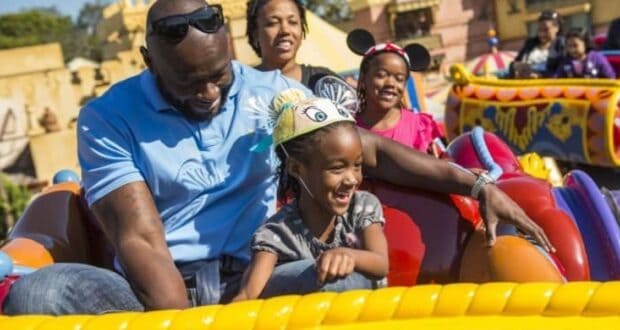 A family enjoys a ride on a colorful amusement park attraction. A father in sunglasses and a blue shirt sits beside his smiling daughter wearing a Disney-themed hat. Other park visitors are visible in the background.