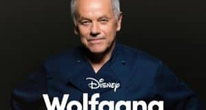 Wolfgang Puck, wearing a dark chef's coat, stands against a dramatic black background with the "Disney Wolfgang" title in front of him, capturing the cinematic flair of a Disney+ food and entertainment documentary.