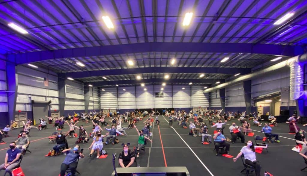 Guests are seated with ample space between each other inside a large gymnasium, carefully following Disney’s signature safety protocols that prioritize everyone’s well-being—just like the attention to detail and guest care you expect in every Disney park experience.