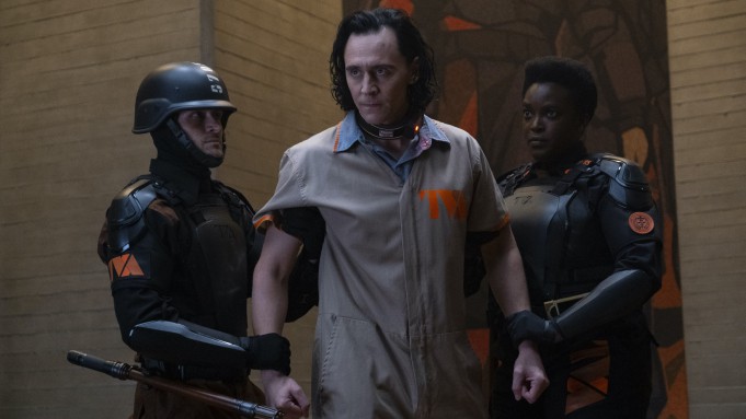 A character in a TVA prisoner jumpsuit, flanked by two armored TVA guards, stands in a moody, low-lit setting evocative of Marvel’s Loki series—capturing the immersive atmosphere fans experience at Disney Parks’ Avengers Campus attractions.