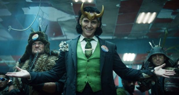 Loki, dressed in his signature green suit and horned helmet, welcomes guests with open arms at a Marvel event, surrounded by fellow costumed characters bringing the Disney+ series to life.