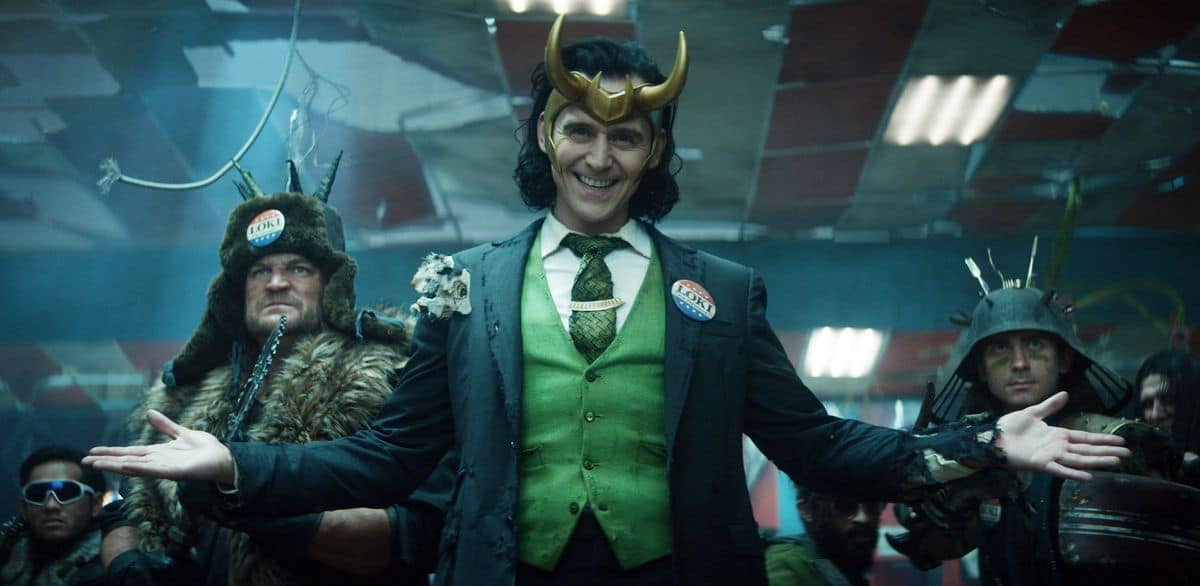Loki, dressed in his signature green suit and horned helmet, welcomes guests with open arms at a Marvel event, surrounded by fellow costumed characters bringing the Disney+ series to life.