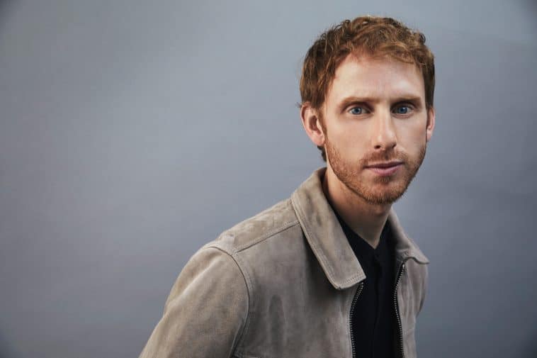 Robert Emms, styled as his character from Star Wars: Andor, stands against a simple gray backdrop in a tan jacket, ready for fans to spot him at a special Star Wars event or themed attraction.