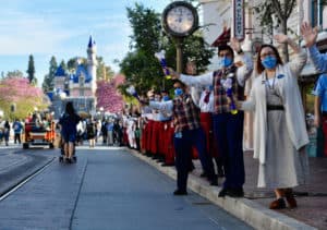 Main Street, U.S.A. comes alive as Disneyland Cast Members sporting masks enthusiastically wave to returning guests in front of Sleeping Beauty Castle; the iconic Main Street clock and vintage trolley complete this heartwarming scene marking the magical reopening of the park.