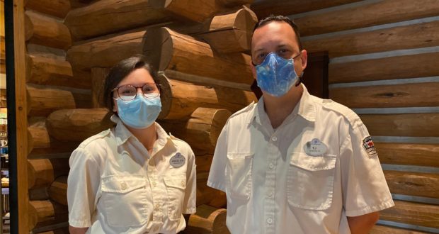Cast Members in crisp white uniforms and masks greet guests in front of the signature log wall at Disney’s Wilderness Lodge, embodying the warm, adventurous spirit of the resort during its grand reopening.