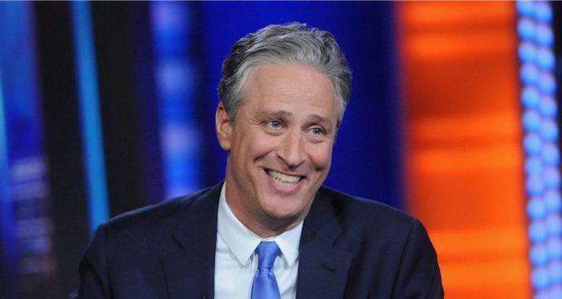 Jon Stewart, dressed in a sharp suit and blue tie, beams as he sits onstage surrounded by vibrant, colorful lights, capturing the excitement and showmanship of a live entertainment spectacular at a top venue.