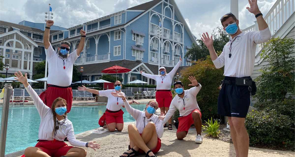 Six Disney lifeguards in signature red shorts and masks pose cheerfully by the Beach Club Resort pool, with the resort’s iconic blue exterior behind them, celebrating the grand reopening as proud Cast Members.