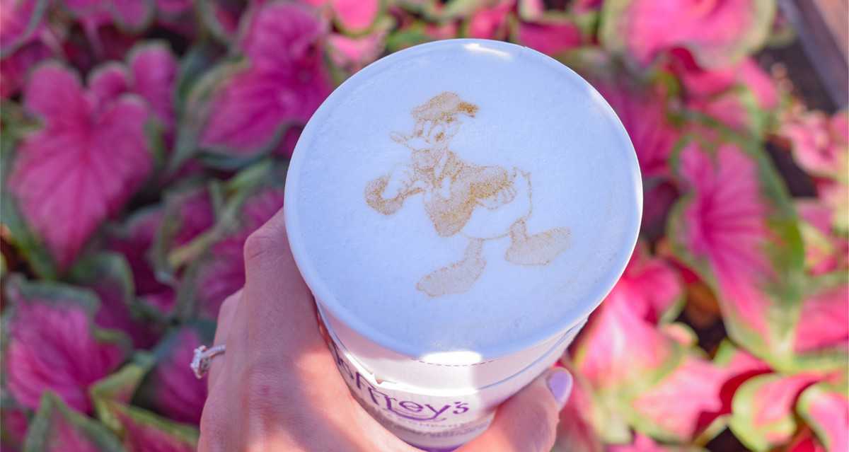 Celebrate Donald Duck’s big day at Disney Springs with a delicious Joffrey’s latte featuring Donald Duck latte art, held up against a dreamy backdrop of blurred pink leaves.