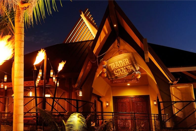 The entrance to Trader Sam's tiki bar glows at night with flickering torches, vibrant tropical decor, and signature wooden doors, inviting Disney fans to experience the magical ambiance unique to both Disneyland and Disney World.