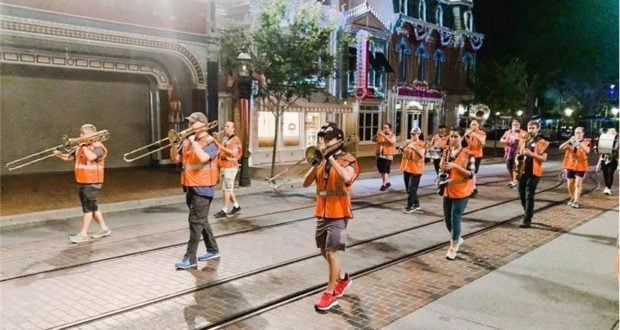 A vibrant marching band in orange vests, echoing the spirited cast members of Disneyland, entertains nighttime crowds as they parade down Main Street, their energetic music adding excitement to the magical park atmosphere.