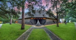 The "Darth Vader House," an eye-catching modern structure with an angular, futuristic roof and expansive windows, sits amidst lush lawns and shade trees, inviting fans of iconic movie architecture to experience a real-life villain’s lair now open for tours or themed events.