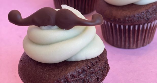 The Mustache Cupcake serves up Disney Springs fun with a chocolate cupcake topped with creamy white frosting and a playful chocolate mustache, all set on a pink background. A sweet treat designed for Father's Day celebrations at Walt Disney World!.
