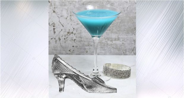 The Glass Slipper Martini—a dazzling blue cocktail served in a martini glass—sits beside a sparkling Cinderella slipper and a shimmering silver bracelet on a pale surface, set against a soft gray backdrop to evoke the enchanting magic of Disney Parks.