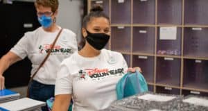 Disney VoluntEARS, wearing masks and signature shirts, organize school supplies during a volunteer event in a backstage storage area.