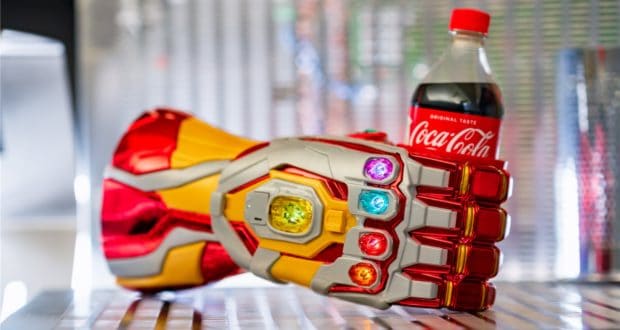 The iconic Infinity Gauntlet, gleaming in red and gold, proudly clutches a Coca-Cola bottle as its vibrant stones shimmer—like you’ve caught Iron Man himself at Avengers Campus, taking a refreshment break between heroic moments.
