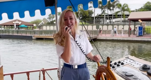 A Disney Watercraft Captain beams with enthusiasm, singing into a microphone as they expertly steer the boat near the dock under sunny skies, capturing the iconic spirit and hospitality of Walt Disney World transportation.