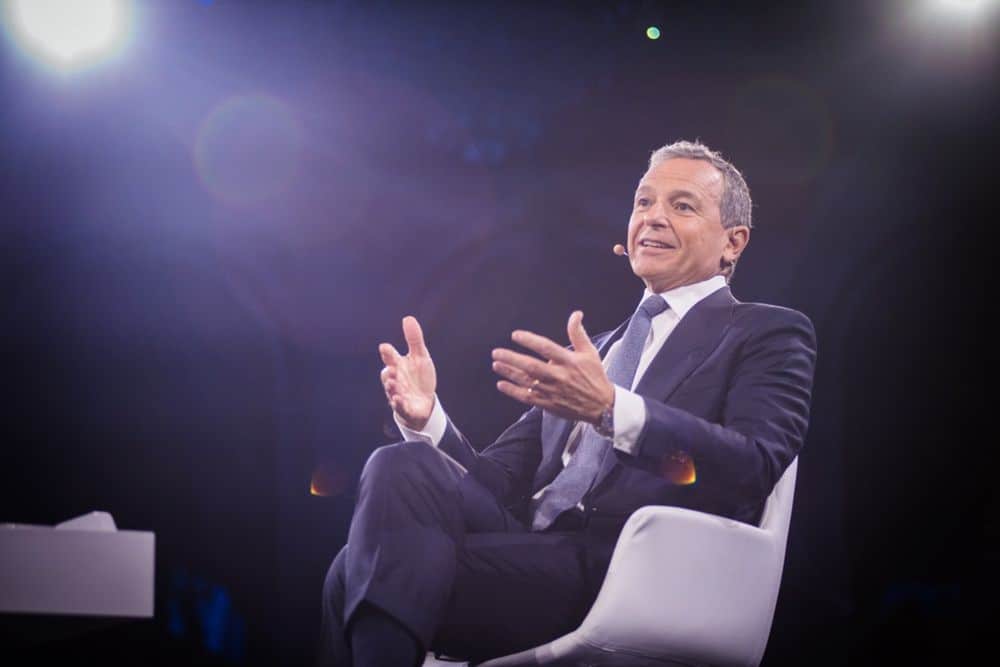 Disney Chairman Bob Iger sits onstage in a suit, animatedly addressing fans and industry guests, with vibrant stage lights illuminating the scene.