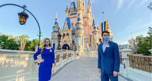 Two friendly Disney Cast Members in matching blue costumes and face masks greet guests in front of Cinderella Castle at Magic Kingdom on a bright, sunny day.