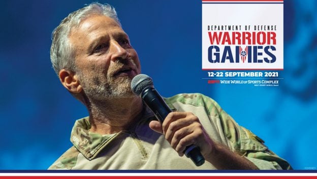 Jon Stewart shares the stage with a service member in camouflage at the DoD Warrior Games, set against a vibrant blue Disney-themed backdrop.