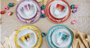 Four vibrant teacup and saucer sets from the Disney Villains dinnerware collection are displayed on an elegantly decorated table, surrounded by flowers, themed napkins, and gold utensils—perfect for fans looking to add a touch of villainous magic to their next Disney-themed gathering or event.