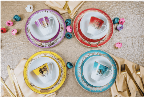 Four vibrant teacup and saucer sets from the Disney Villains dinnerware collection are displayed on an elegantly decorated table, surrounded by flowers, themed napkins, and gold utensils—perfect for fans looking to add a touch of villainous magic to their next Disney-themed gathering or event.