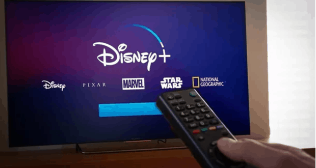 A Disney fan readies the remote in front of a TV showcasing Disney+ with vibrant Disney, Pixar, Marvel, Star Wars, and NatGeo logos—bringing the magic of Main Street U.S.A., Avengers Campus, and Galaxy’s Edge right into their living room as they prepare to stream the latest weekly episode.