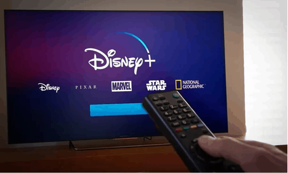 A Disney fan readies the remote in front of a TV showcasing Disney+ with vibrant Disney, Pixar, Marvel, Star Wars, and NatGeo logos—bringing the magic of Main Street U.S.A., Avengers Campus, and Galaxy’s Edge right into their living room as they prepare to stream the latest weekly episode.