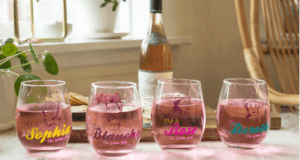 Four glasses of pink cocktails, each tagged with a Golden Girl’s name, are displayed in front of a wine bottle and lush greenery—inviting Epcot festival-goers and Disney+ viewers alike to raise a glass in celebration of The Golden Girls.
