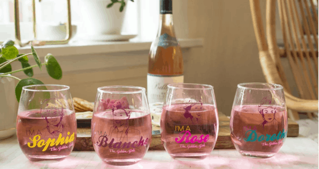 Four glasses of pink cocktails, each tagged with a Golden Girl’s name, are displayed in front of a wine bottle and lush greenery—inviting Epcot festival-goers and Disney+ viewers alike to raise a glass in celebration of The Golden Girls.