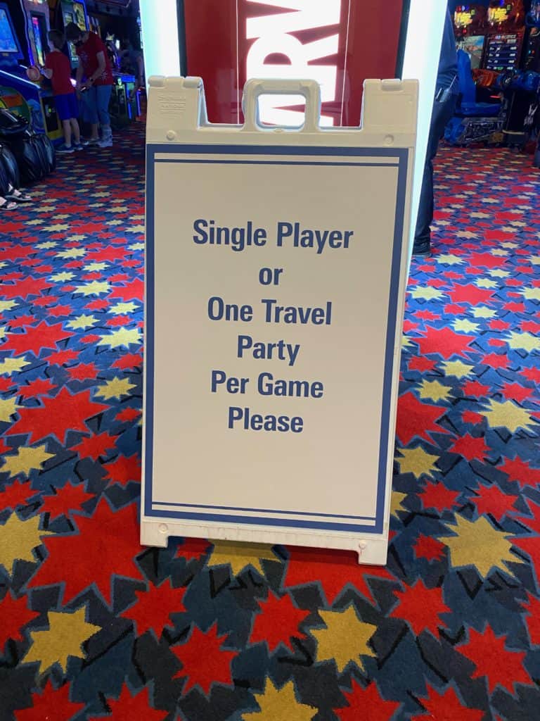 A sign placed on a vibrant arcade carpet at Disney World reads: "Single Player or One Travel Party Per Game Please"—a friendly reminder seen throughout Disney World arcades and playgrounds to help guests enjoy safe, magical fun together.