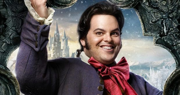 LeFou, dressed in his signature purple coat and red bow tie, beams as he poses with a decorative frame in front of a snow-dusted castle backdrop, capturing the whimsical charm of Beauty and the Beast at a Disney park.