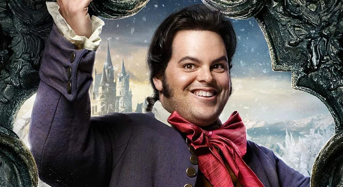 LeFou, dressed in his signature purple coat and red bow tie, beams as he poses with a decorative frame in front of a snow-dusted castle backdrop, capturing the whimsical charm of Beauty and the Beast at a Disney park.