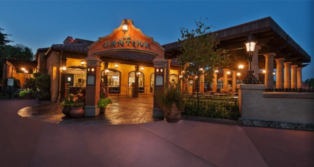 The warmly illuminated exterior of a Mexican-themed quick-service restaurant at Walt Disney World, featuring an inviting arched entrance and spacious outdoor seating, welcomes guests at dusk.