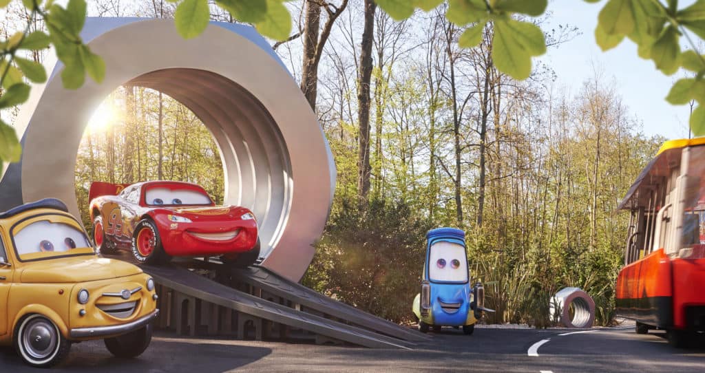 Smiling Cars characters line up near a circular ramp in the sun-drenched outdoor Cars Road Trip attraction at Disneyland Paris, surrounded by trees and celebrating the park’s latest magical addition.
