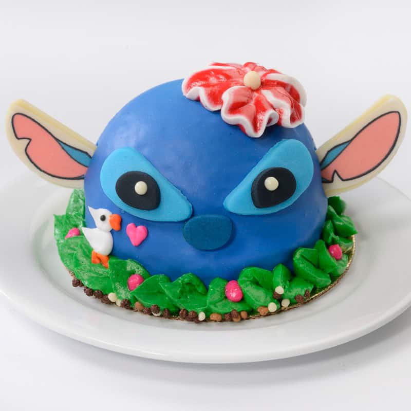 Stitch-themed blue dome cake with a red flower, signature ears, and bird decoration, served on a white plate with swirls of green icing—a whimsical Stitch Treats dessert inspired by Disney’s Polynesian Village Resort, available for a limited time to Disney fans.