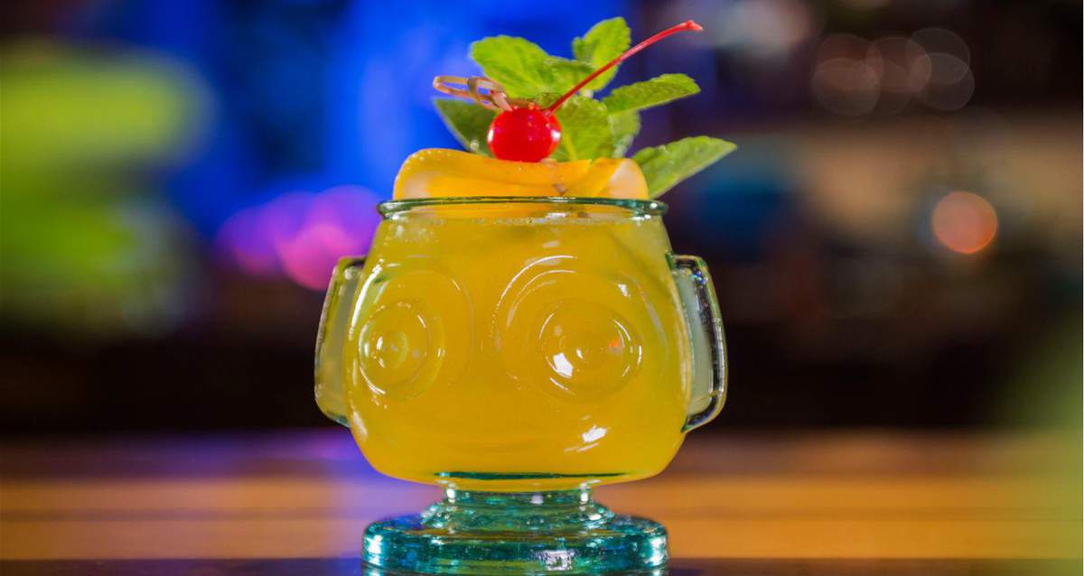 A vibrant yellow cocktail served in a quirky tiki-style glass, topped with a cherry, orange slice, and fresh mint, sits on a wooden surface—just like the fun tropical drinks you’d sip at Trader Sam’s in Disney World or Disneyland.