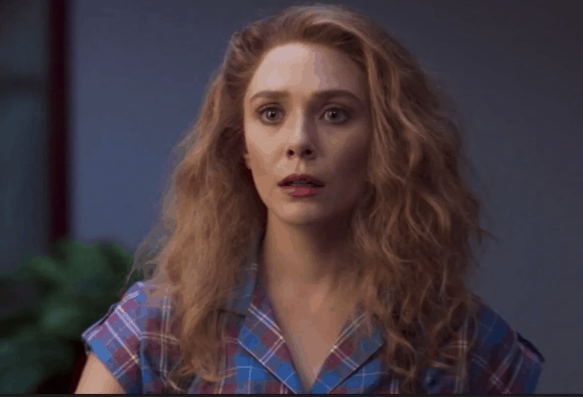 A guest with wavy red hair in a blue plaid shirt stands indoors at Avengers Campus, looking genuinely surprised with a plant in the background—like she just learned that WandaVision Season 2 could be coming to Disney+.