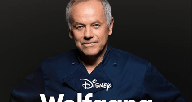 Wolfgang Puck stands confidently in a navy chef's coat with arms folded beneath the "Disney Wolfgang" title on a sleek black background, capturing his star power as a culinary legend and inviting Disney fans to discover the stories behind his remarkable journey.