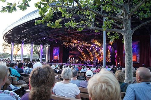 Fans gather at EPCOT’s Food & Wine Festival, soaking up live music from the Entertainment Lineup on a covered stage nestled among lush trees in the afternoon sun.