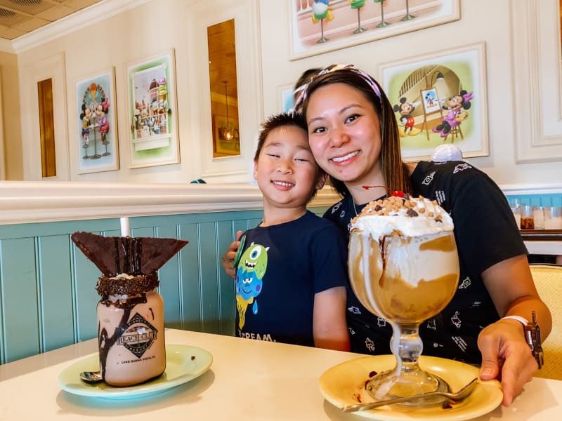 A mom and her child enjoy massive chocolate desserts at a whimsically decorated cafe by Crescent Lake, the ideal sweet stop along your Walt Disney World dessert crawl.