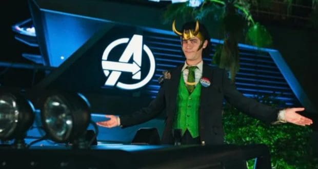 President Loki greets fans with open arms in front of the glowing Avengers logo at night, perfectly channeling the playful chaos of Avengers Campus.