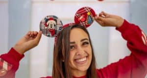 A guest at Disneyland beams with holiday cheer in a red sweater and a festive Holiday Knit Ear Headband inspired by Spirit Jersey, playfully holding the cozy knit ears with both hands.