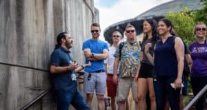 A lively tour host entertains a crowd of visitors under the sun near a concrete wall, captivating them with fascinating stories about the site’s dramatic ties to defense history, much like an immersive experience in a themed attraction.
